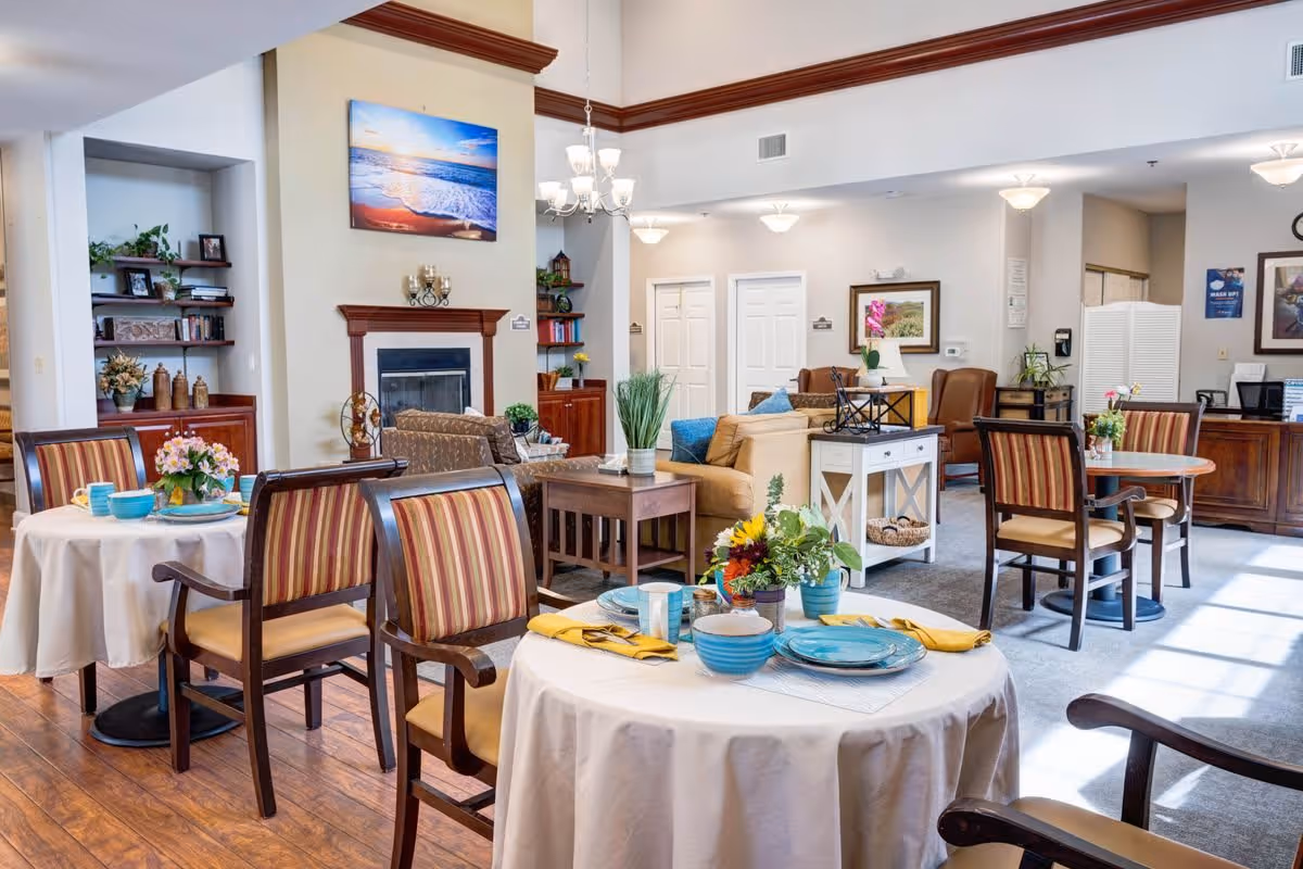 A bright and welcoming common area in a senior living facility featuring round tables with striped cushioned chairs, set with blue plates, bowls, and yellow napkins. The room has a fireplace with a beach-themed painting above it, a chandelier, and comfortable seating including sofas and armchairs. Shelves with decorative items and plants are visible, along with a reception desk in the background.