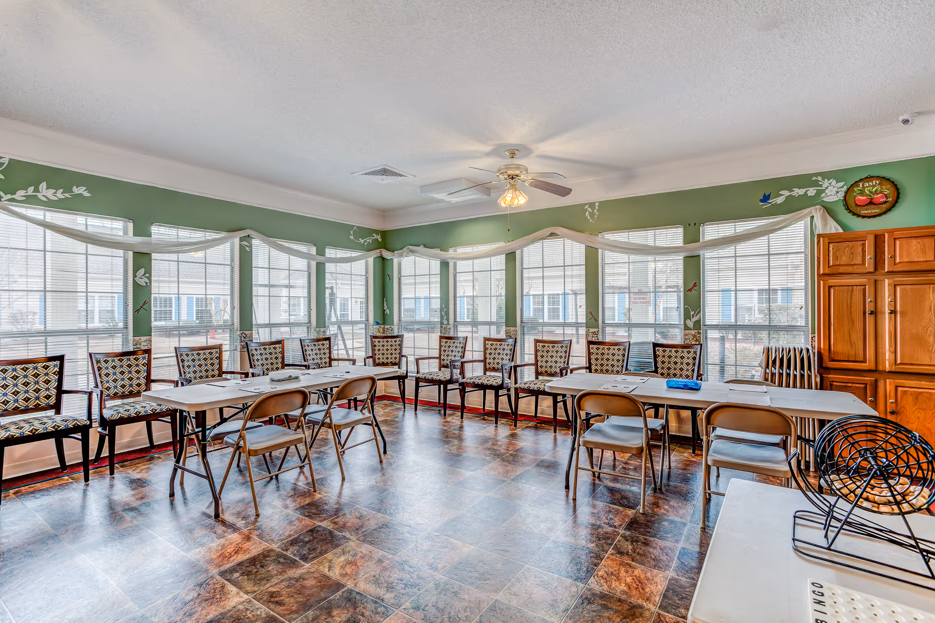 A bright room with large windows along two walls, green walls with white decorative accents, and a ceiling fan with lights. The room contains two long tables arranged in an L-shape with several folding chairs and patterned armchairs around them. There is a wooden cabinet on the right side and a bingo cage on a small table in the foreground.