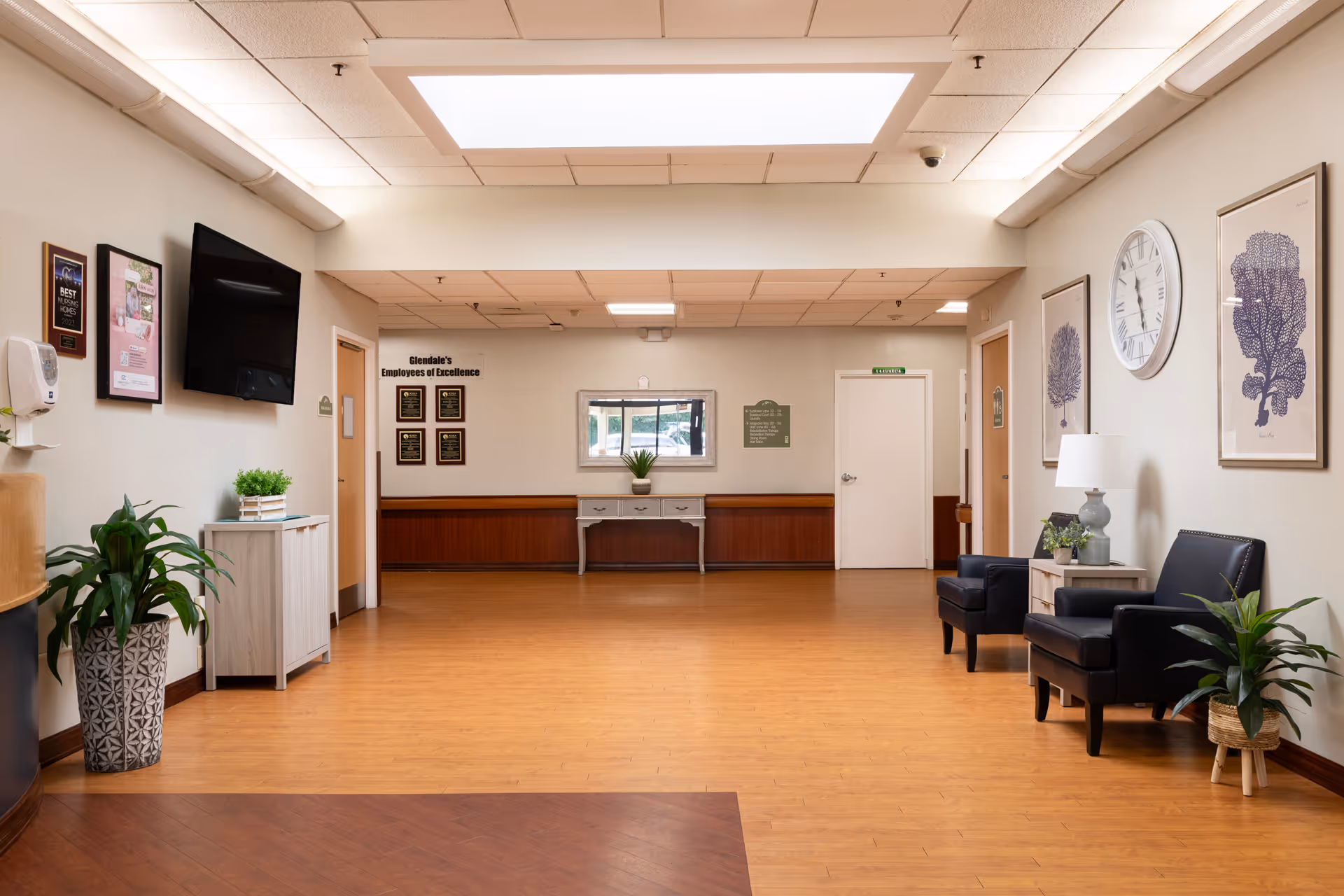 Bright, well-lit reception lounge with seating, plants, wall clock, TV and award plaques.