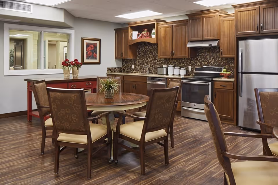 Communal dining area with a round wooden table and chairs in front of a fully equipped kitchen with stainless steel appliances and wood cabinets.