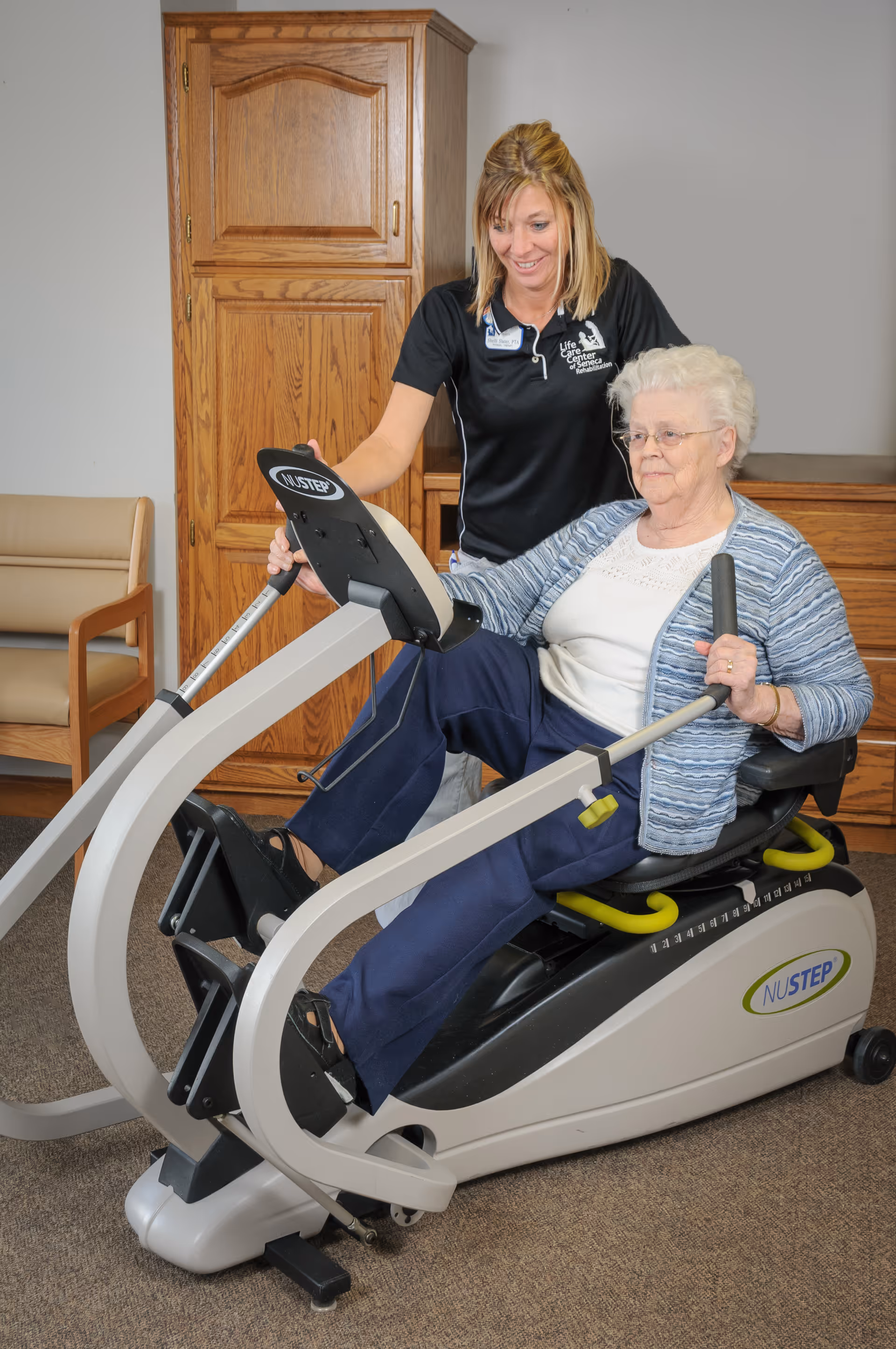An elderly woman using a NuStep exercise machine while being assisted by a female staff member wearing a Life Care Center of Seneca shirt in a room with wooden cabinets and chairs.