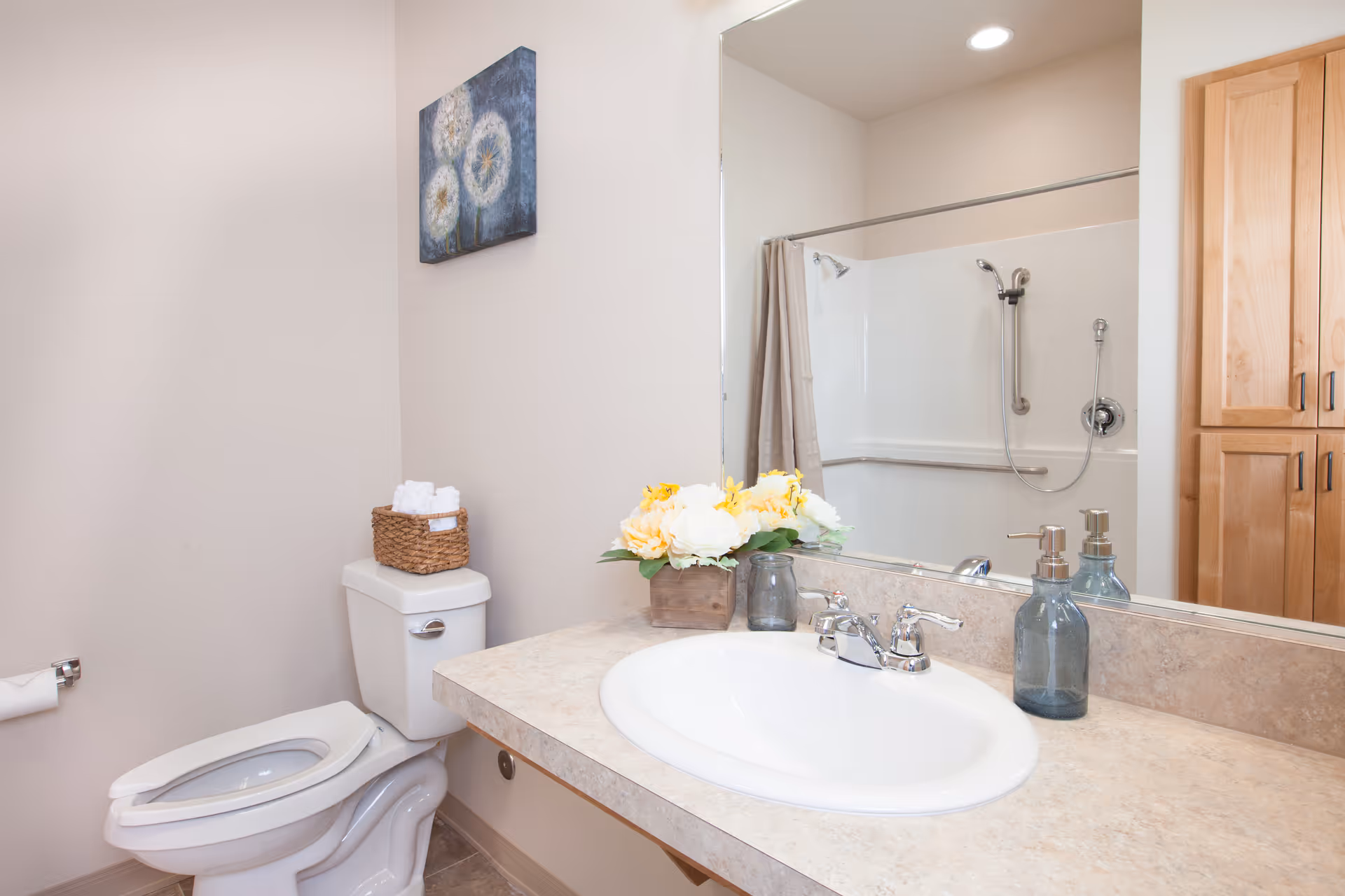 A clean bathroom featuring a white toilet with a basket of rolled towels on top, a countertop with a white sink, a soap dispenser, a small vase with yellow and white flowers, a large mirror, and a shower with a curtain and grab bars. There is also a wooden cabinet visible in the reflection of the mirror.