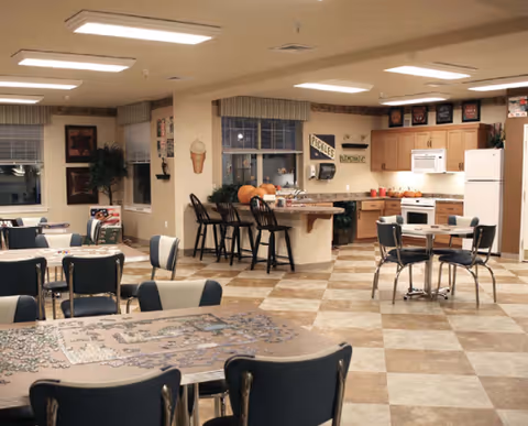 A spacious common area with a kitchen and dining space. The kitchen has wooden cabinets, a microwave, stove, and a countertop with bar stools. Several tables and chairs are arranged around the room, with one table featuring a partially completed jigsaw puzzle. The room is well-lit with ceiling lights and decorated with wall art and plants.