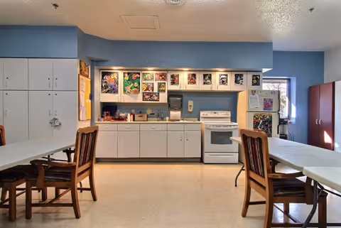 Communal dining area with tables and chairs facing a small kitchenette with cabinets, sink, stove and colorful artwork on the back wall.