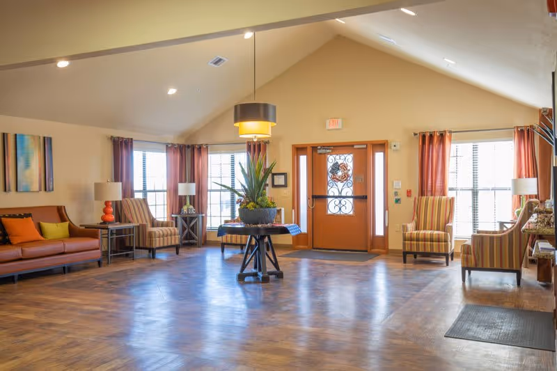 Spacious, well-lit lobby with seating, striped armchairs, a central plant on a round table, and an orange entrance door.