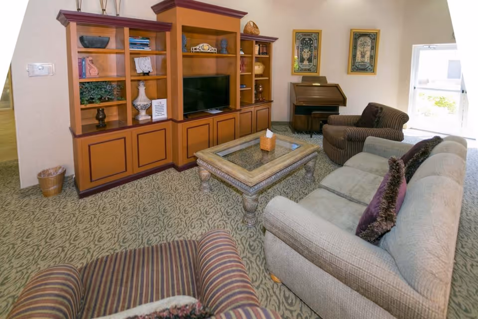 Light-filled common living room with sofas and armchairs arranged around a decorative coffee table and built-in wooden shelving with a TV.