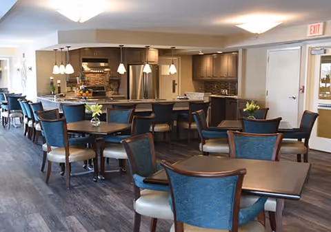 A spacious dining area with multiple square and round tables surrounded by blue upholstered chairs with wooden frames. The room features a modern kitchen in the background with stainless steel appliances, pendant lighting, and a countertop with bar stools. The flooring is dark wood, and the walls are light-colored, creating a bright and welcoming atmosphere.