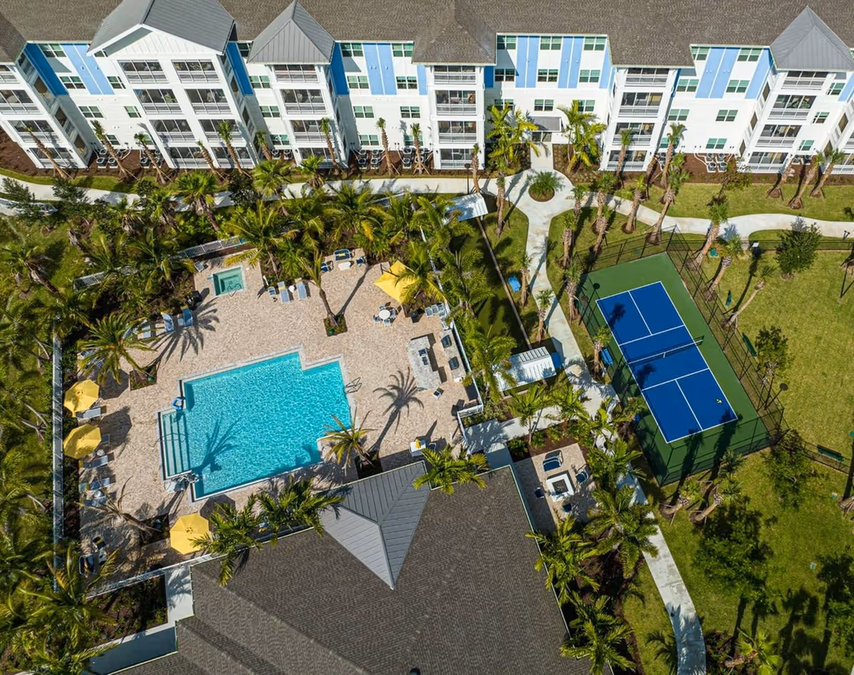 Aerial view of an outdoor courtyard with a swimming pool, lounge areas, palm trees, and a tennis court beside a multi-story residential building.