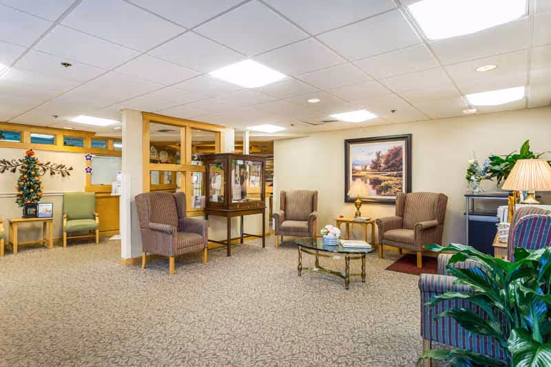 A bright common lounge in Sterling Care with upholstered armchairs around a glass coffee table, plants, and framed artwork.
