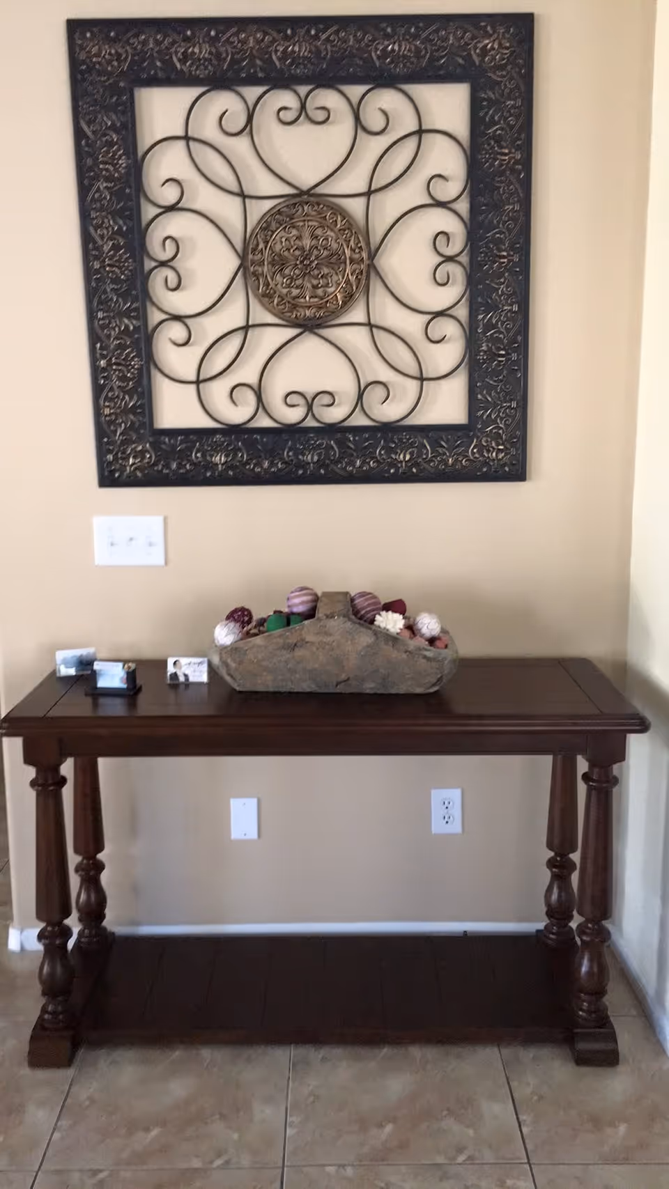 Wooden console table against a beige wall with ornate metal wall art above and a decorative bowl of potpourri on top.