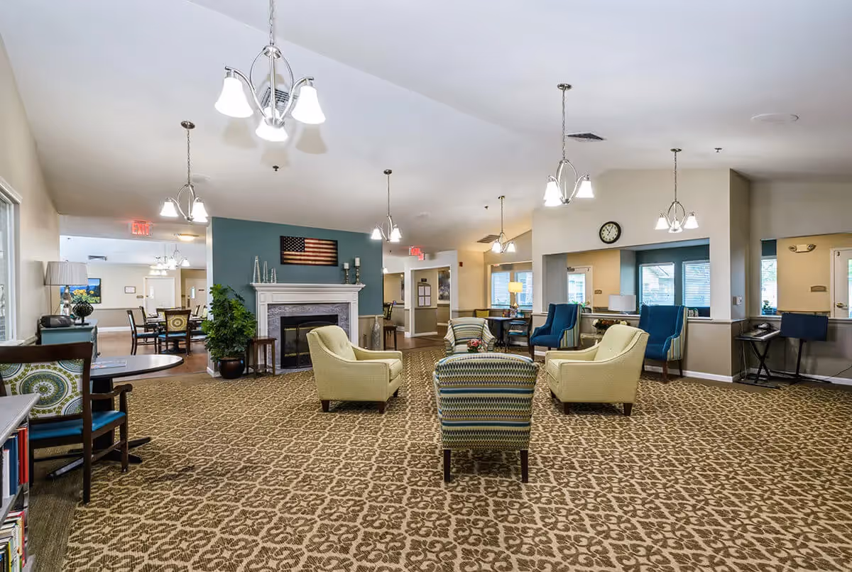 A spacious senior living facility common area with patterned carpet, multiple armchairs arranged around a central ottoman, a fireplace with an American flag above it, and several hanging light fixtures. The room has a cozy and welcoming atmosphere with a mix of blue and beige tones, and a dining area is visible in the background.