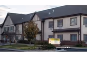 Exterior view of a two-story assisted living facility with Tudor-style architecture, featuring beige walls with dark wooden trim, multiple windows, a small landscaped area with a tree, and a sign in front of the building.