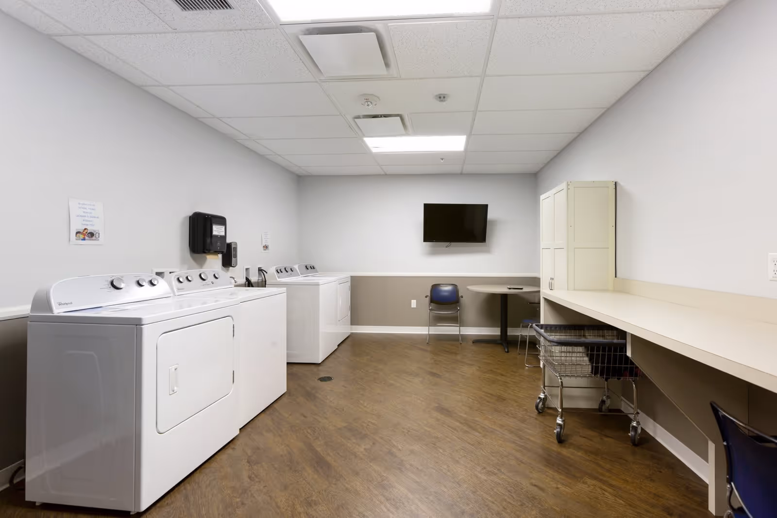 A laundry room with two washing machines and two dryers along the left wall. There is a long counter with a laundry cart underneath on the right side, a small table with a chair, a wall-mounted TV, and a storage cabinet against the far wall. The room has wood flooring and white walls with a beige wainscoting.