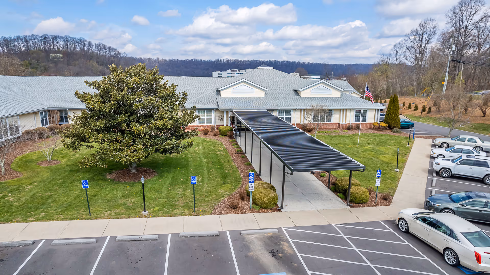 Exterior view of American House Bristol facility showing a single-story building with a covered walkway entrance, green lawn with trees and bushes, several handicap parking spaces, and parked cars. The sky is partly cloudy with hills in the background.