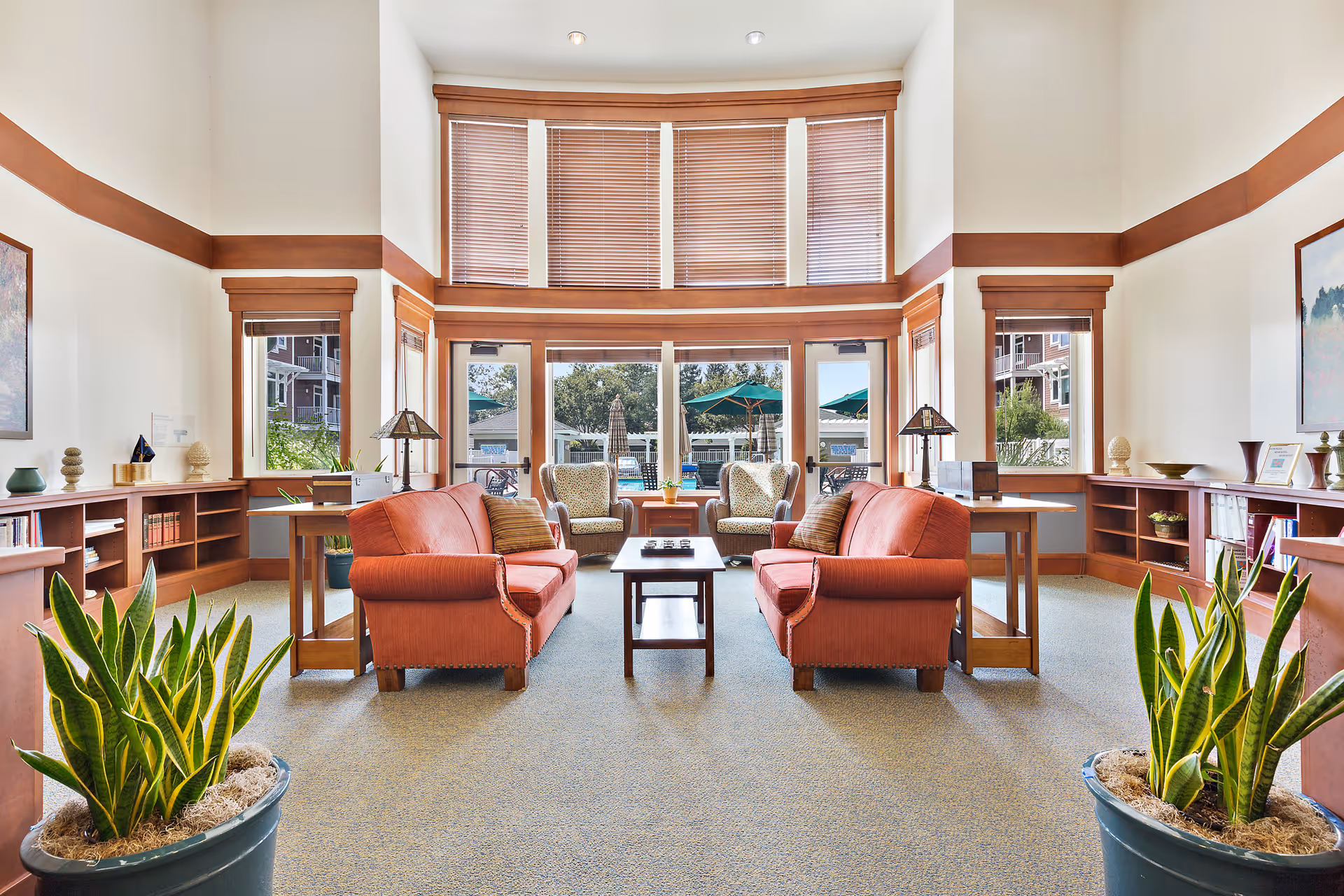 A bright and spacious living room area with two orange sofas facing each other across a wooden coffee table. Behind the sofas are two armchairs positioned near large windows with wooden blinds, allowing natural light to fill the room. The room features wooden trim accents, built-in bookshelves along the walls, two potted plants in the foreground, and decorative lamps on side tables next to the sofas. Outside the windows, green umbrellas and outdoor seating are visible.
