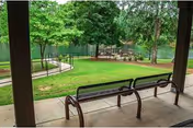 View from a covered patio area with two metal benches overlooking a green lawn with trees, a paved walkway, and an outdoor seating area with tables and chairs in the background.