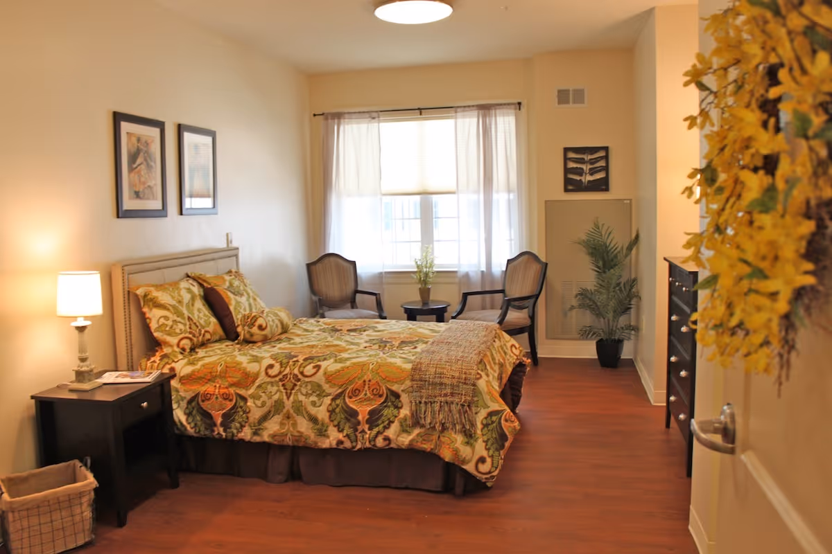 A furnished bedroom with a patterned bed, nightstand and lamp, two chairs by a window, and a dresser on wood floors.