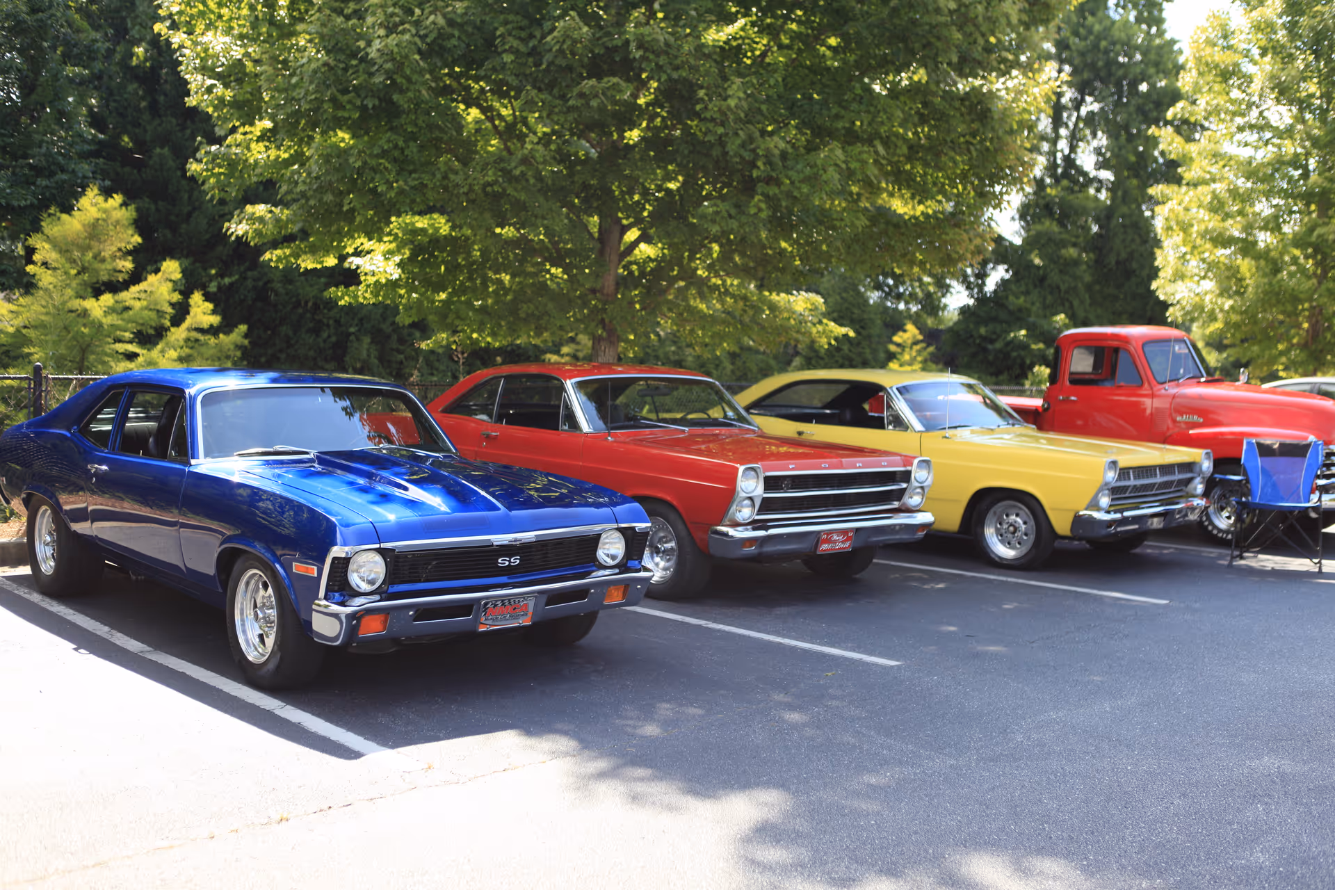 Four classic vehicles parked in a row in an outdoor parking lot, including a blue Chevrolet SS, a red Ford, a yellow car, and a red vintage pickup truck, with trees and greenery in the background.