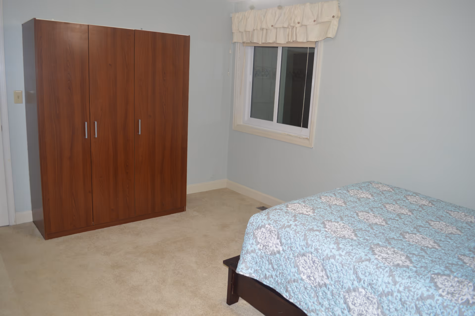 Simple bedroom with a wooden wardrobe, a bed covered by a blue patterned quilt, and a window with a valance.