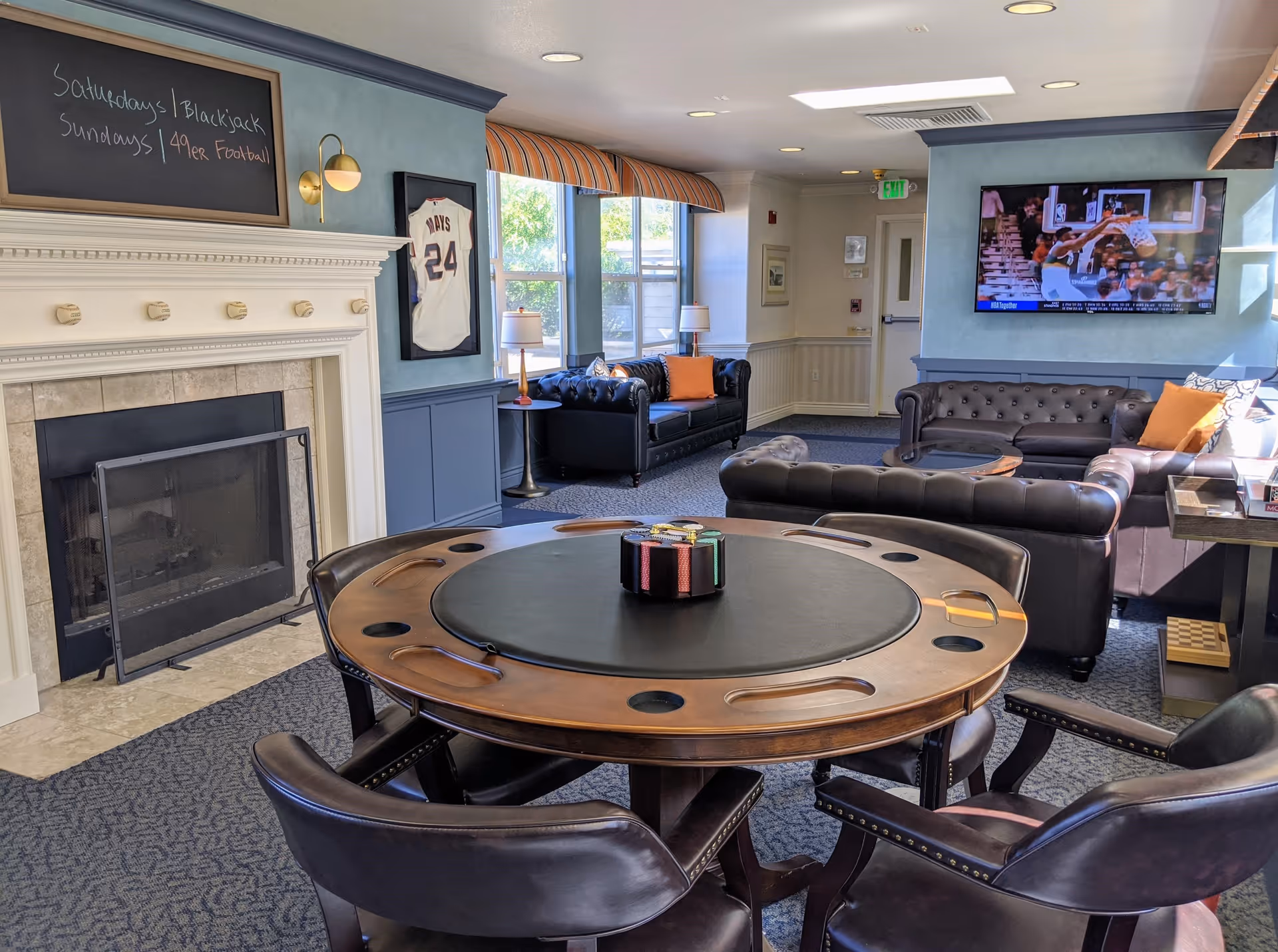 A cozy common area with a round poker table and four leather chairs in the foreground. Behind the table, there are two leather sofas with orange and patterned pillows, a fireplace with a white mantel, a framed sports jersey on the wall, and a flat-screen TV mounted on the wall showing a basketball game. The room has blue walls, carpeted floor, and large windows letting in natural light.