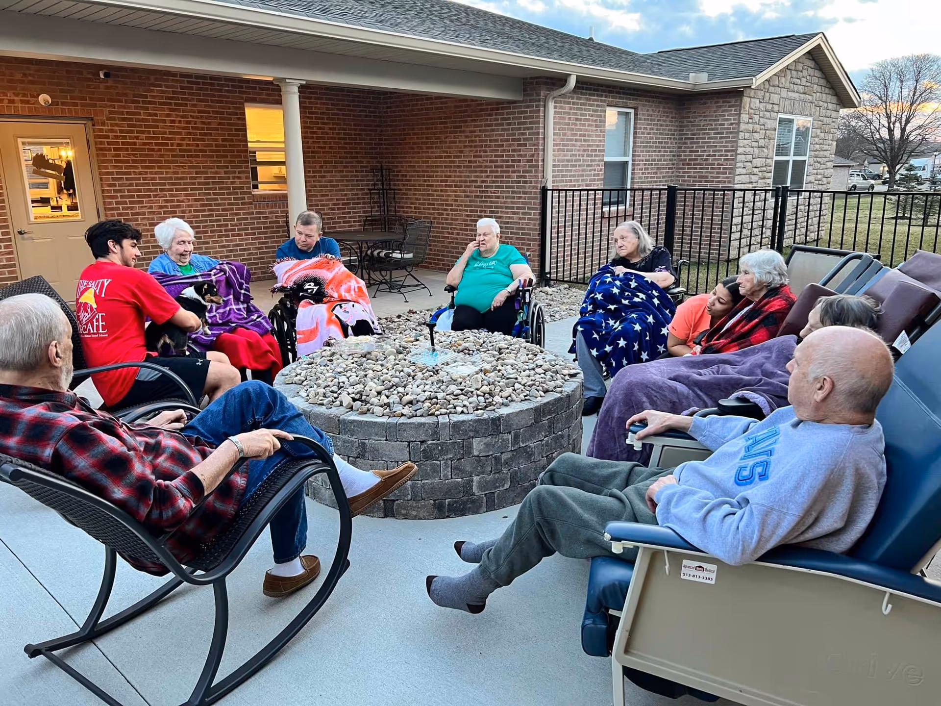 A group of elderly people and two caregivers sitting around a circular stone fire pit on an outdoor patio at a senior living facility. Some of the elderly individuals are in wheelchairs and others are seated in chairs, many wrapped in blankets. The background shows a brick building and a fenced yard under a partly cloudy sky.
