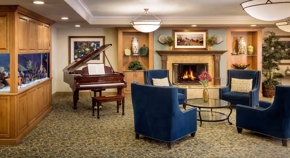 A cozy living room area in Atria Santa Clarita featuring a grand piano with sheet music, a built-in aquarium, a fireplace with a painting above it, and four blue upholstered armchairs arranged around a glass coffee table with a vase of flowers.