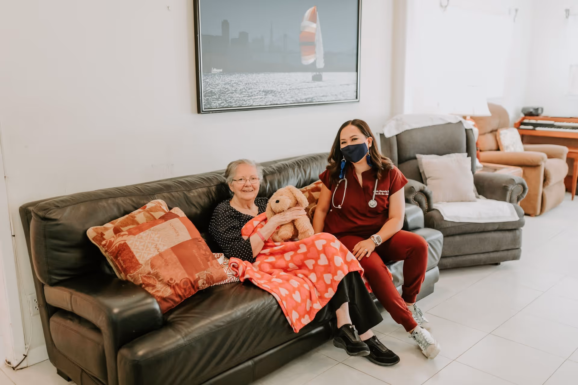 An elderly woman sitting on a black leather couch covered with a red blanket with heart patterns, holding a stuffed dog toy. Next to her is a healthcare worker wearing a maroon uniform and a black face mask, sitting on the couch armrest. The room has a gray armchair, a brown recliner, a piano, and a framed picture of a sailboat on the wall.