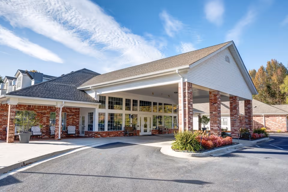 Exterior view of Bridgewood Gardens senior living facility showing a covered entrance with brick pillars, large windows, and a driveway under a blue sky with some clouds. There are potted plants and outdoor seating near the entrance.