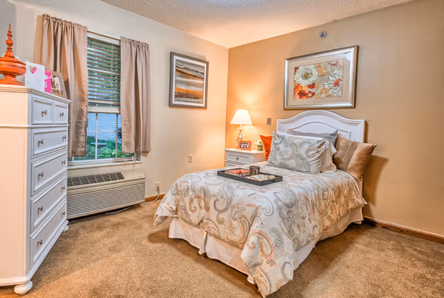 A cozy single bedroom with a patterned twin bed, nightstand and lamp, white dresser, window with curtains, and framed artwork on beige walls.