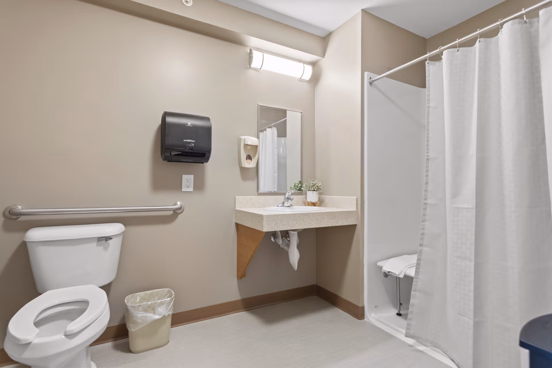 A clean and accessible bathroom with a toilet, a wall-mounted grab bar, a small trash can, a sink with a mirror above it, a soap dispenser, and a shower area with a white curtain and a foldable shower seat.