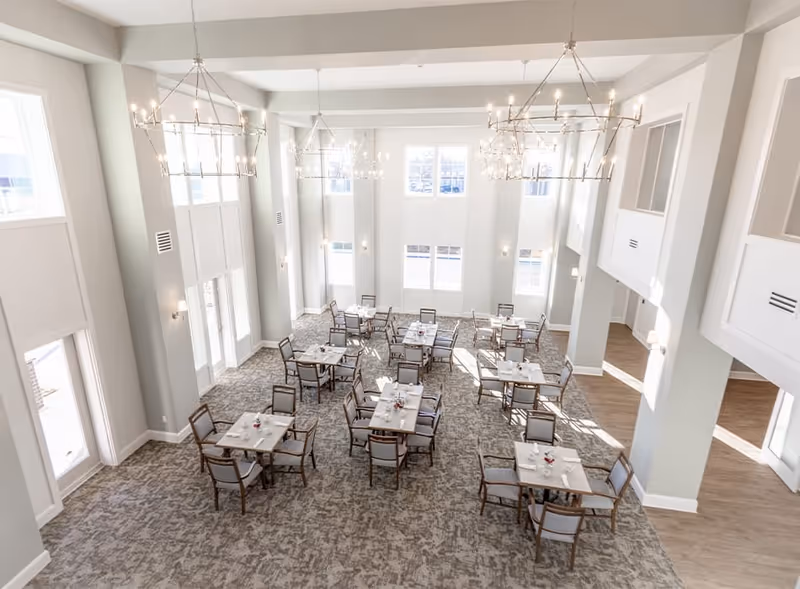 Bright and spacious dining room with multiple square tables and chairs arranged neatly on a patterned carpet. Large windows allow natural light to fill the room, and elegant chandeliers hang from the high ceiling.