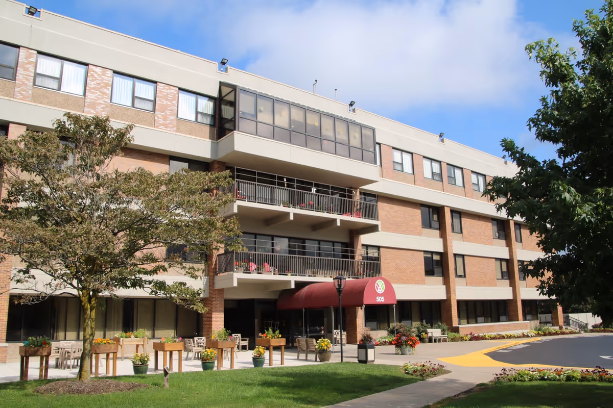 Exterior view of a multi-story senior living facility building with balconies, a red awning over the entrance, trees, and flower pots along the sidewalk under a partly cloudy sky.