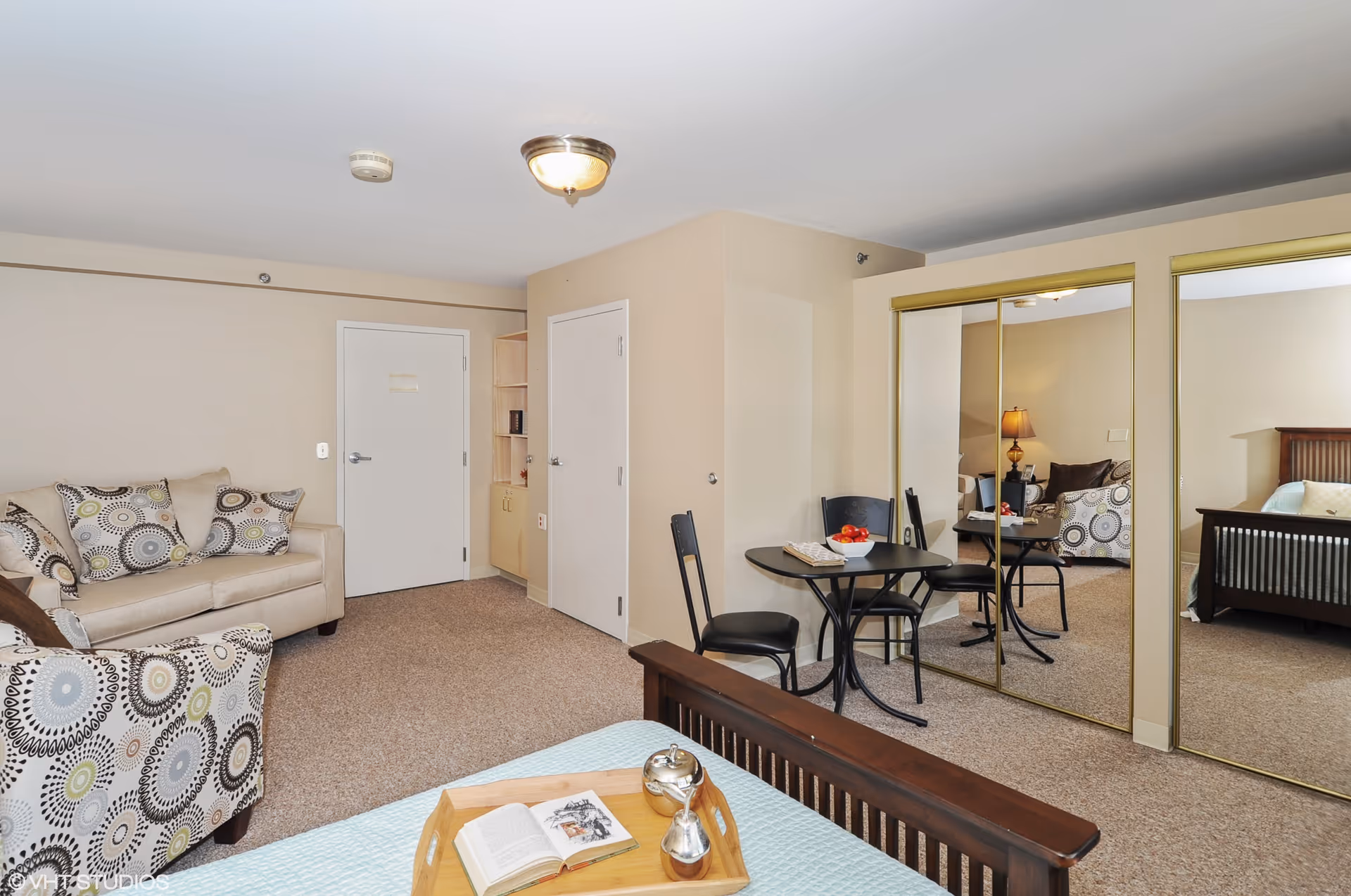 A cozy living space with a beige sofa and patterned armchair, a small round table with two black chairs, a bed with a wooden headboard, and mirrored sliding closet doors reflecting the room. The walls are light beige, and the floor is carpeted.