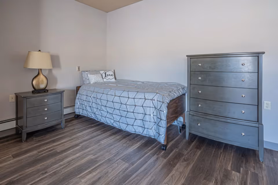 A simple bedroom with a single bed covered in a gray patterned comforter and pillows, including one with the word 'Love'. Next to the bed is a small nightstand with a lamp, and on the right side of the bed is a tall dresser. The room has wood flooring and plain light-colored walls.