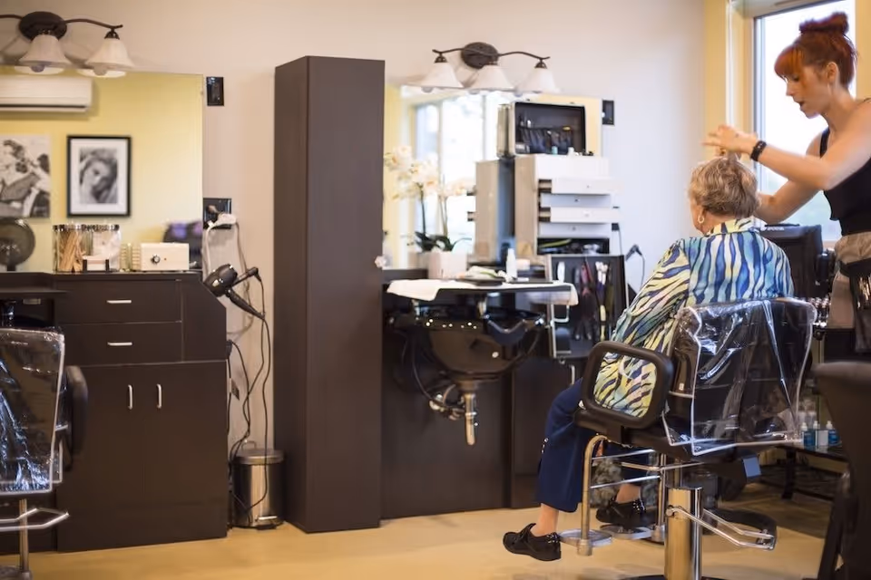 A hair salon area inside a senior living facility where a stylist is cutting the hair of an elderly woman seated in a salon chair covered with a protective cape. The room has salon equipment, mirrors, and cabinets with hair care tools.