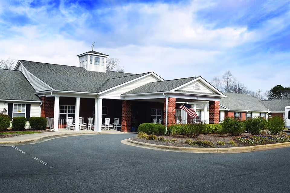 Exterior view of a single-story senior living facility building with a covered entrance supported by white columns and brick pillars. The building has a gray shingled roof with a small cupola on top. There are white rocking chairs on the porch and an American flag displayed near the entrance. Shrubs and landscaped plants surround the driveway and entrance area under a partly cloudy blue sky.
