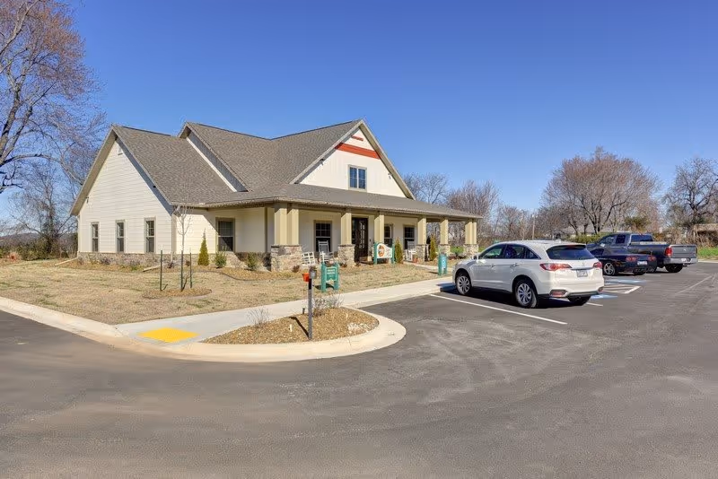 Exterior view of a single-story building with a gabled roof and a covered porch, surrounded by a parking lot with several parked vehicles and leafless trees under a clear blue sky.