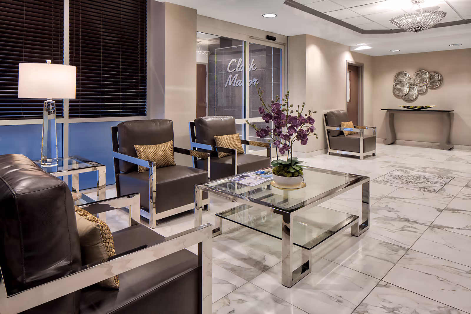 A modern and elegant waiting or lounge area with black leather chairs featuring chrome armrests, a glass coffee table with a purple orchid centerpiece, a side table with a lamp, marble flooring, and a wall with the words 'Clark Manor' visible through glass doors.