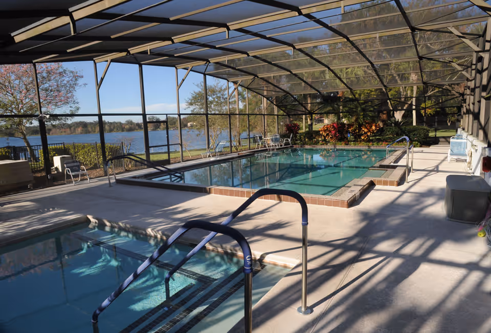 Indoor swimming pool area with a smaller pool or hot tub in the foreground and a larger pool in the background, enclosed by a screened structure. There are metal handrails for pool entry, several chairs around the pool, and a view of a lake and trees outside the enclosure.