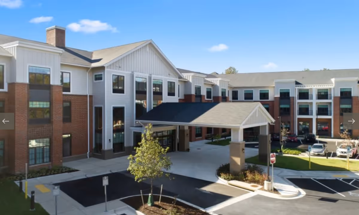 Exterior view of a senior living facility named Elancé at Tuckahoe, showing a multi-story building with a covered entrance, parking spaces, and a small landscaped area with a tree under a clear blue sky.
