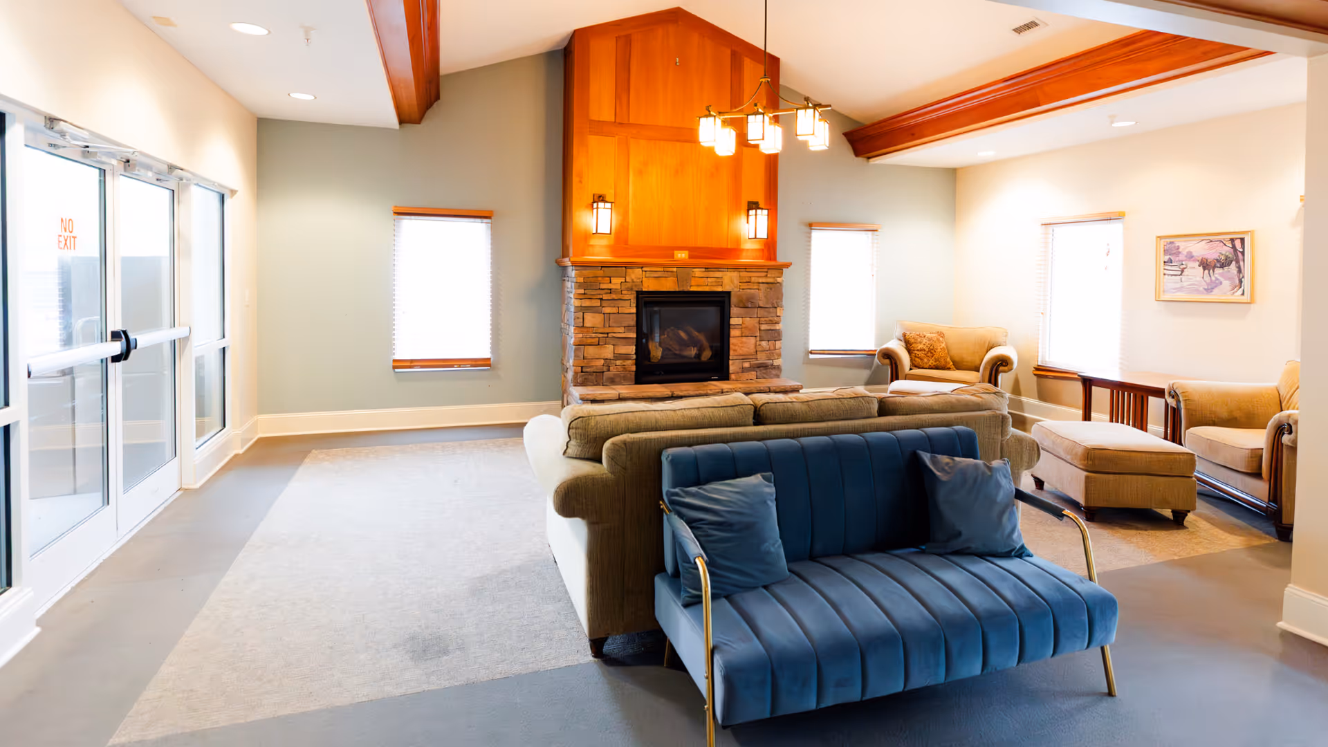 A bright and spacious living room area with a stone fireplace centered on a wooden accent wall. The room features a blue velvet sofa with two pillows in the foreground, a beige sofa behind it, and two beige armchairs with an ottoman near the windows. There are three windows with blinds, a wooden ceiling beam, and a modern chandelier hanging from the ceiling. Double glass doors with a 'No Exit' sign are visible on the left side.