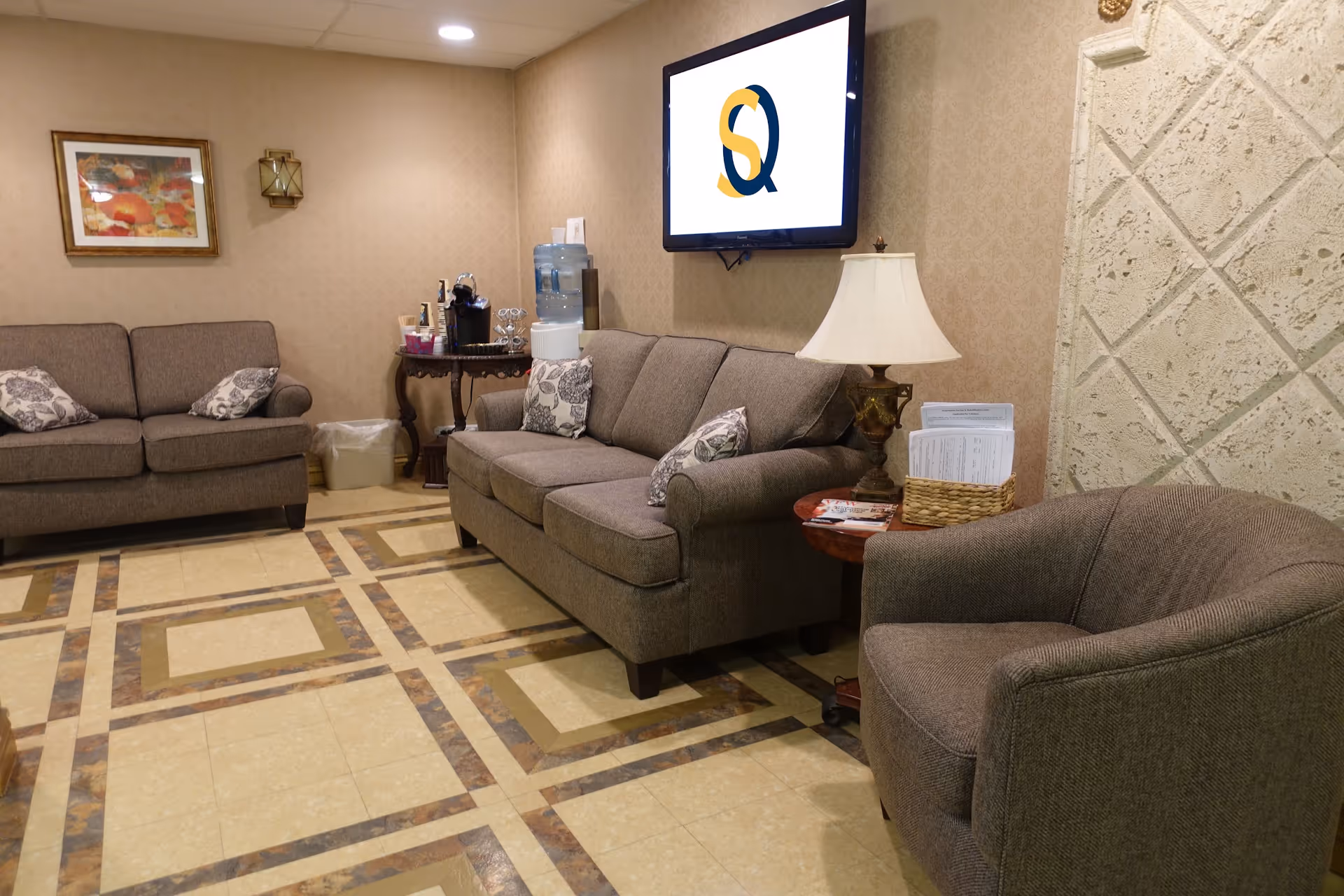 A cozy sitting area in Susquehanna Nursing & Rehabilitation Center featuring two brown sofas and a matching armchair with patterned cushions. A small wooden table with a lamp and a basket of papers is beside the armchair. A water dispenser and coffee station are on a small table against the wall. A flat-screen TV mounted on the wall displays a logo with intertwined letters 'S' and 'Q'. The room has beige patterned wallpaper, a framed floral picture, and a tiled floor with a geometric design.