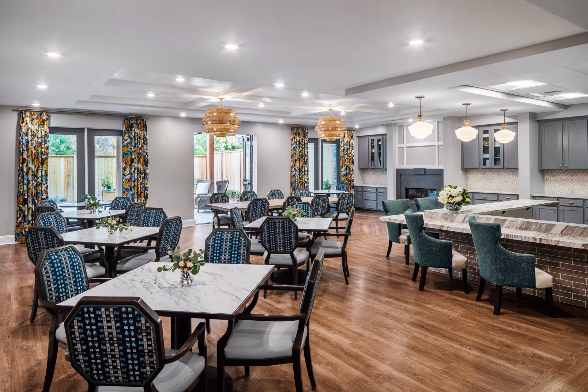 A spacious and well-lit dining area with multiple tables and chairs arranged neatly. The tables have marble tops and small floral centerpieces. There are large windows with colorful floral curtains allowing natural light to enter. The room features a kitchen area with a marble countertop bar and green upholstered bar stools. The floor is wooden, and the ceiling has recessed lighting and decorative light fixtures.