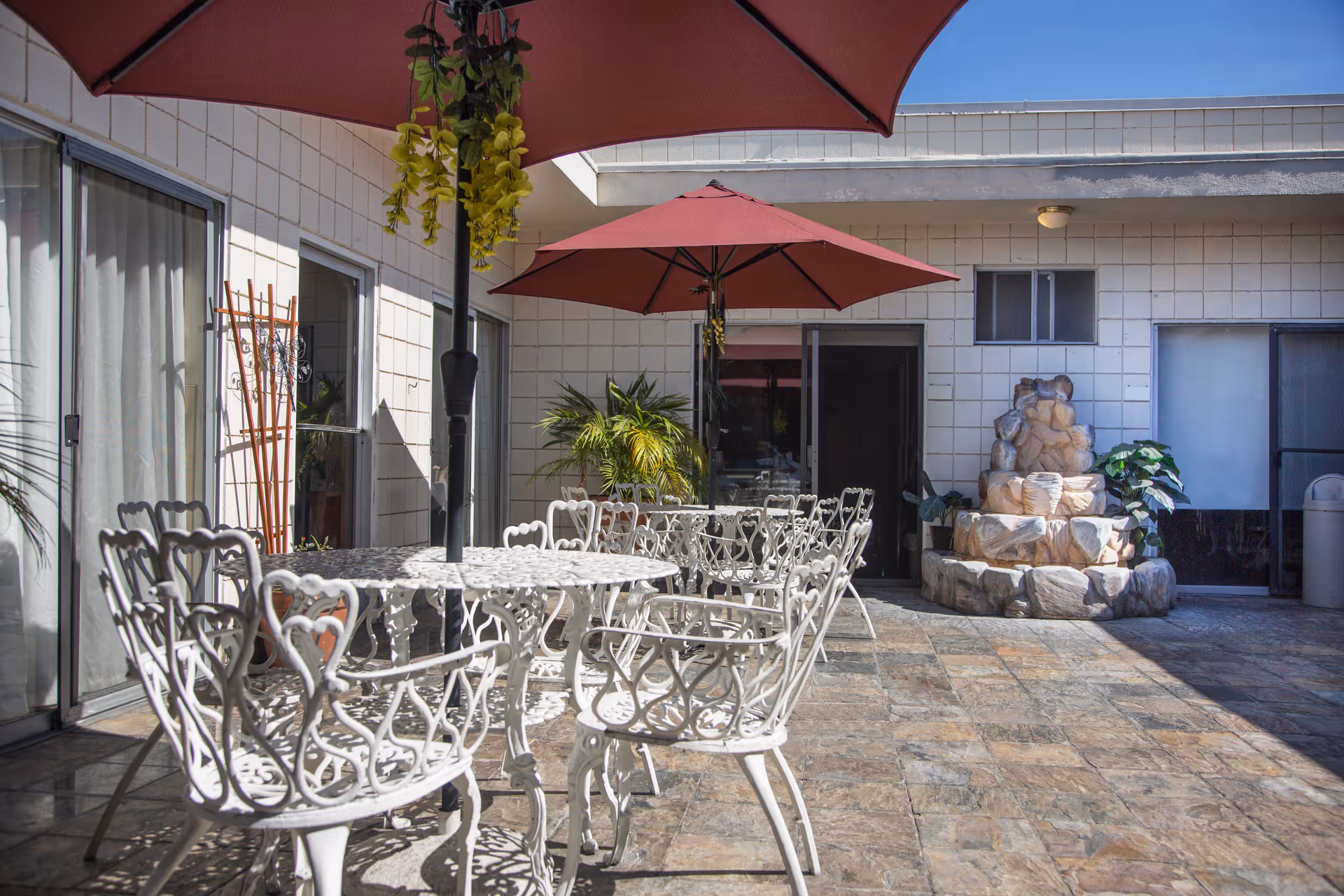 Outdoor patio area with white metal tables and chairs, red umbrellas providing shade, potted plants, and a stone water fountain against a tiled building wall.
