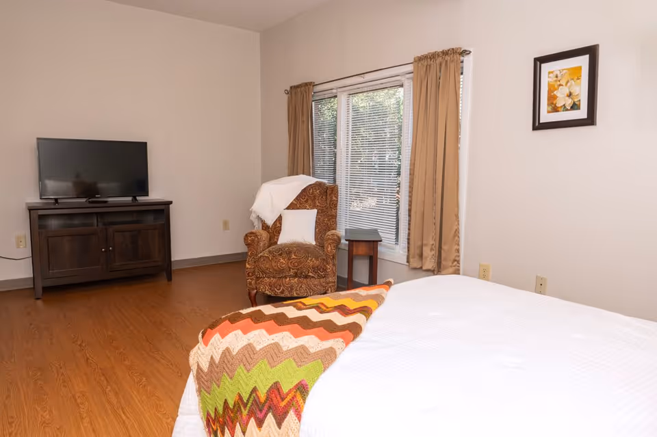 A bedroom in an assisted living facility featuring a bed with a colorful zigzag patterned blanket, a brown patterned armchair with a white pillow and throw, a small wooden side table, a window with beige curtains and blinds, a flat-screen TV on a dark wooden cabinet, and a framed floral painting on the wall.