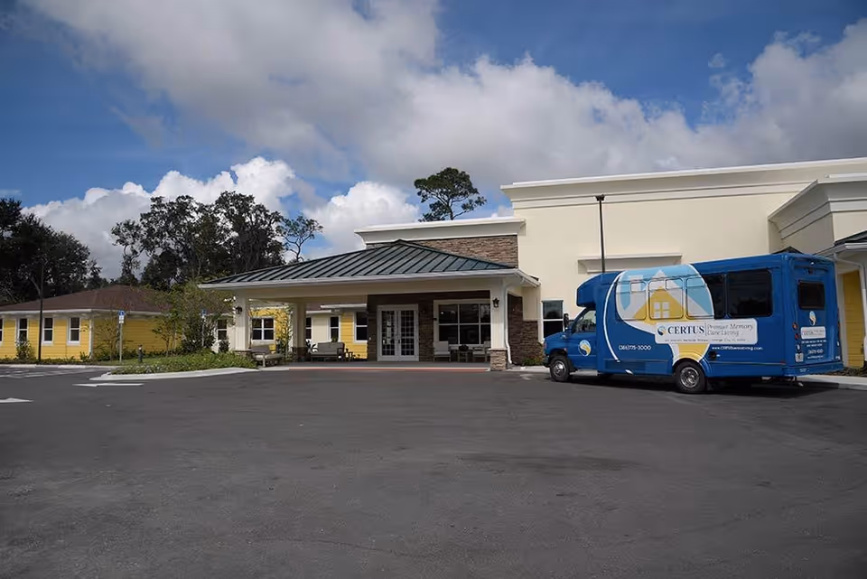 Blue CERTUS memory care bus parked in front of beige building entrance with a covered canopy