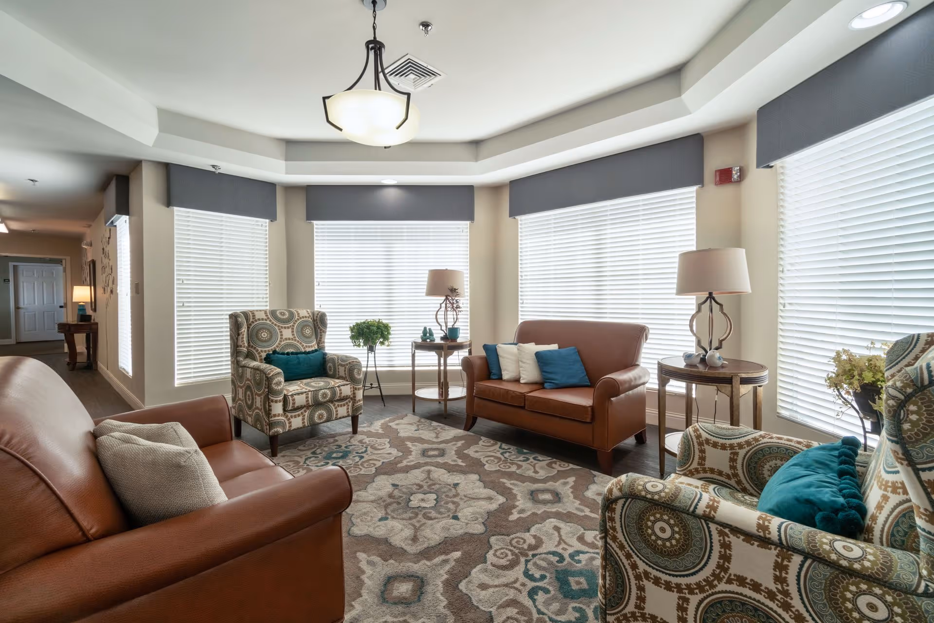 A bright and cozy senior living common area with large windows covered by white blinds and gray valances. The room features two brown leather sofas with decorative pillows, two patterned armchairs with teal cushions, a patterned area rug, two side tables with lamps, and a small plant on a stand near the windows.