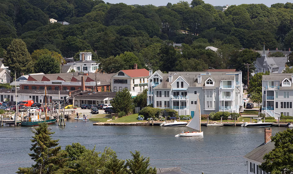 View of a waterfront area with residential buildings and boats docked along the shore. A sailboat is sailing on the water, and there are trees and greenery in the background.