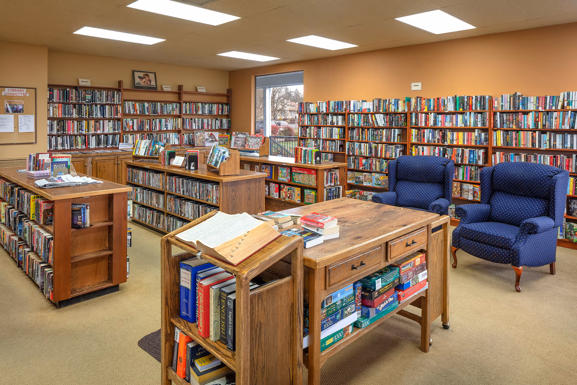A cozy library room with wooden bookshelves, tables, stacked books and two blue upholstered armchairs.