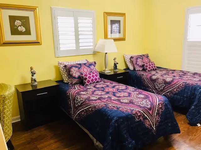 A bedroom with two twin beds featuring colorful purple and pink patterned bedding. Each bed has matching pillows and is separated by a dark wooden nightstand with a white lamp and small decorative figurines. The walls are painted yellow and decorated with framed artwork. There are white plantation shutters on the windows, and the floor is wooden.